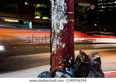 Wallpaper of black trash bags near a worn red pole with car trail lights on the background at night in the street. No people.