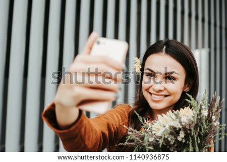 A picture of woman taking selfie with flowers. She holds them in hand. Girl looks at phone and smiles. She is happy. Isolated on striped and white background
