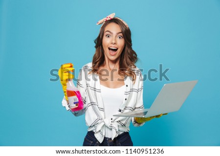 Portrait of european happy housewife 20s in yellow rubber gloves holding detergent sprayer and laptop during cleaning isolated over blue background