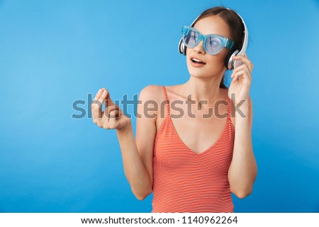 Portrait of an attractive young girl in swimsuit wearing sunglasses and listening to music with headphones isolated over blue background