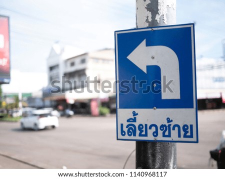 Blue rectangle traffic sign "Turn left" in Thai language with a background of road in a city.