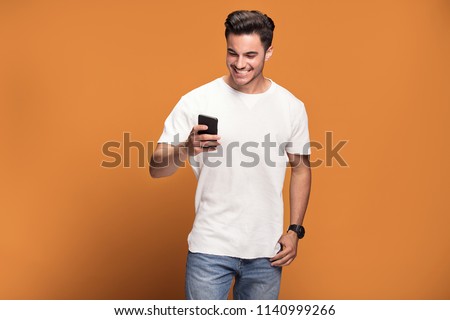 Photo of happy handsome man holding mobile phone in hand, texting. Young guy with big toothy smile posing on yellow background wearing white t-shirt.