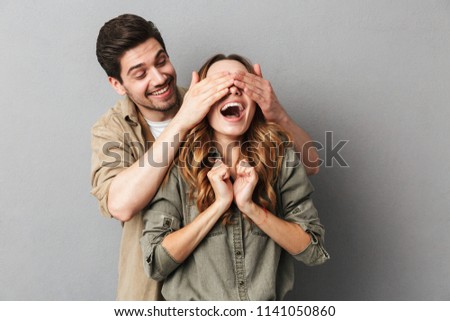 Portrait of a happy young couple, man covering his girlfriends eyes isolated over gray background Royalty-Free Stock Photo #1141050860