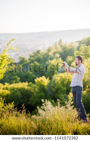 A man in a shirt and trousers takes pictures of himself and nature on the phone. Portrait of a man at sunset against the backdrop of the mountains that makes a selfie