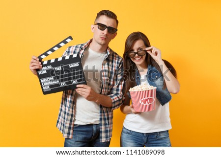 Young smiling couple woman man in 3d glasses watching movie film on date holding classic black film making clapperboard bucket of popcorn isolated on yellow background. Emotions in cinema concept