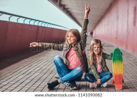 Portrait of two little sisters dressed in trendy clothes sitting together on a skateboard at bridge footway.