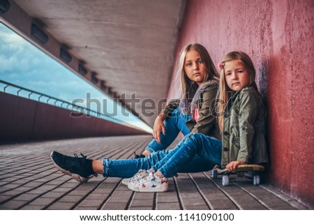 Portrait of two little sisters dressed in trendy clothes leaning on a wall while sitting on a skateboard at a bridge footway.