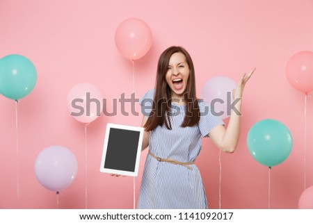 Portrait of crazy young woman in blue dress holding tablet pc computer with blank empty screen spreading hands screaming on pink background with colorful air balloons. Birthday holiday party concept
