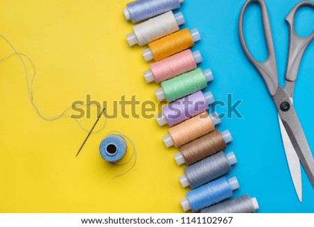Vertical row of coils with colored threads, scissors,  needle and  bobbin on  bright yellow-blue background.