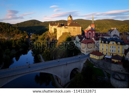Aerial view on gothic Castle Loket, Burg Elbogen, surrounded by river with bridge against blue sky and green hills,massive fortification illuminated by setting sun.Close to Karlovy vary,Czech republic