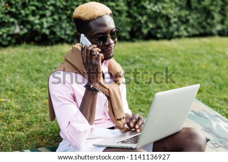 Young american man speak on the phone while sitting on the green grass with laptop