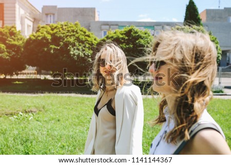 Two stylish hipster girls in glasses are walking in the street. Bright summer photoshoot for women. Beautiful sisters enjoying sun outdoors.
