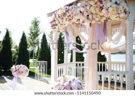 Outdoor Wedding. Ceremony. Wedding arch. Arch made from wood decorated with pastel cotton textile, purple, beige and white flowers and greenery on bank of river