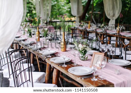 Wedding. Banquet. The festive table for guests, decorated with a composition of white and pink flowers and greenery, there are candles, served with crockery in wooden summer house. Royalty-Free Stock Photo #1141150508
