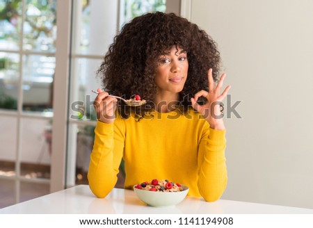 African american woman eating cereals, raspberries and blueberries doing ok sign with fingers, excellent symbol