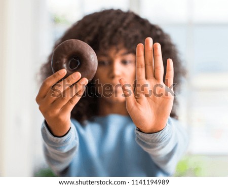 Young african american woman holding chocolate donut at home with open hand doing stop sign with serious and confident expression, defense gesture
