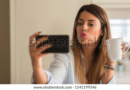 Beautiful young woman taking a selfie with phone while drinking cup of coffee at home.