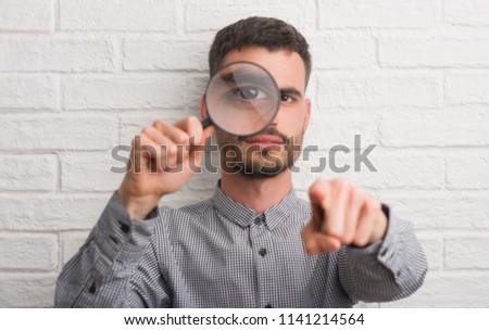 Young adult man over brick wall using magnifying glass pointing with finger to the camera and to you, hand sign, positive and confident gesture from the front