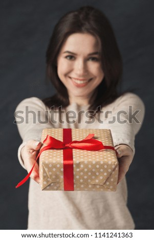Picture of young brunette woman holding gift, posing and looking at camera over grey background.