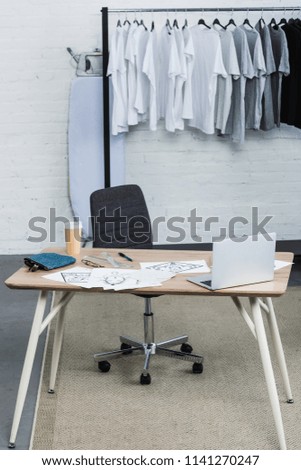 selective focus of t-shirts on hangers and working table with laptop and paintings in clothing design studio