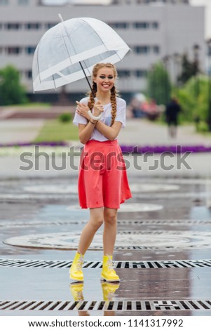Young pretty girl with two braids in yellow boots and with transparent umbrella stands near fountain. Rainy day in city.