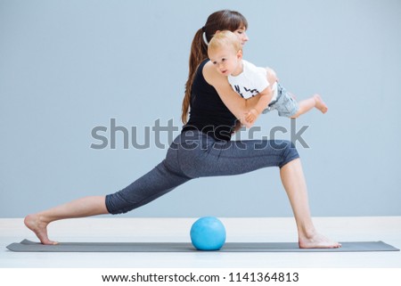 young sporty mother does physical yoga or pilates exercises together with her toddler baby son over gray background. Fitness, happy maternity and healthy lifestyle concept.