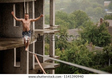 Brave man walking on a slackline and raising hands up against the empty building on rainy day