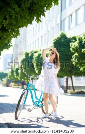 Female is standing on a city street looking up holding a vintage bicycle and a hat on her head. Lightness of movement, blond dress, sunglasses attached to the charm of photography