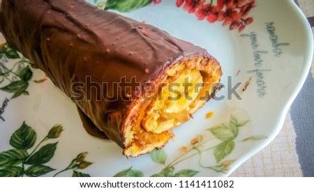 A photo of a homemade Swiss roll, also known as jelly roll, or cream roll served on a floral dish. The roll is like a sponge cake  filled with banana, condense milk, biscuits, dough, and chocolate.