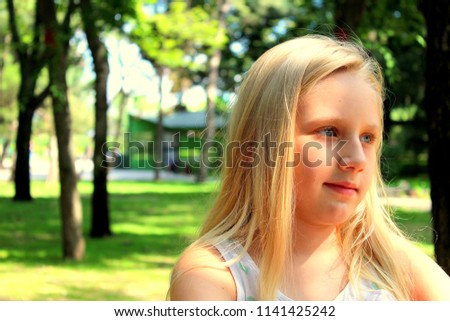 A young blonde girl with long hair is in a green park and shows emotions.