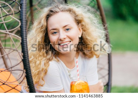 Horizontal shot of pretty smiling European female has wavy bushy light hair, holds fresh cocktail, sits on hanging chair, breathes fresh air. Positive emotions, recreation and people concept