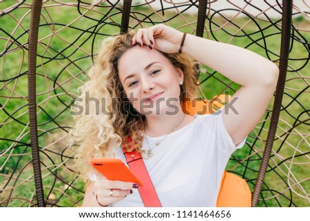 Beautiful delighted female blogger with curly light hair, uses smart phone for posting new posts on her web site, rests in hanging chair, uses wireless internet connection. People, recreation concept