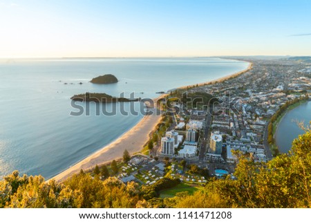 Mount Maunganui stretches out below as sun rises on horizon and falls across ocean beach and buildings below Royalty-Free Stock Photo #1141471208
