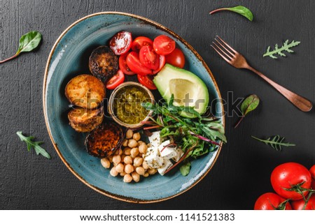 Healthy Buddha bowl dish with avocado, tomato, cheese, chickpea, fresh arugula salad, baked potatoes and sauce pesto in black background. Dieting food, clean eating, top view, flat lay, copy space