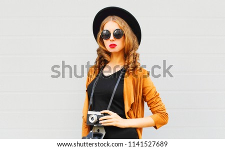 Cool girl model with retro film camera wearing elegant hat, brown jacket posing outdoors over city grey background
