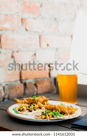 portrait of a soft fried shrimp taco with corn, salad and sauce on a white plate on a wooden table with a pint of beer in the background