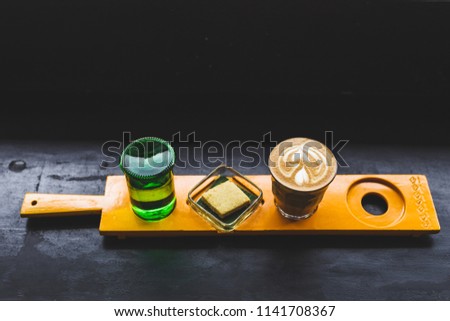 Unusual serving coffee in a cafe: cappuccino, water and biscuit on yellow wooden stand on dark background