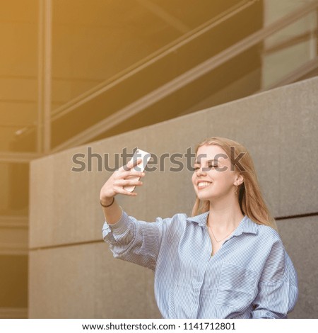 Shot of a laughing young woman  taking a selfie while standing outside near the grey wall, sunny summer day.