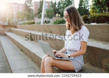 Beautiful girl is working and studying behind a laptop in the park at sunset
