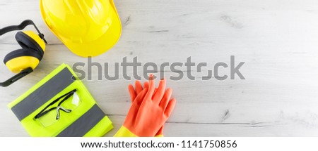 Banner image of construction safety gear including a hard hat, folded reflective jacket, goggles, and rubber gloves flat lay on white wooden surface with copy space Royalty-Free Stock Photo #1141750856