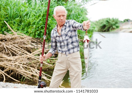 Cheerful adult fisherman removing fish from hook at lakeside on summer day