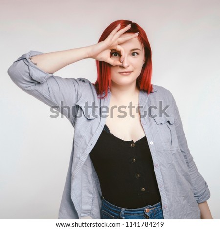 Red haired plus size ordinary woman standing on a neutral grey background. Emotional portrait. She holds an okay gesture in front of her eye