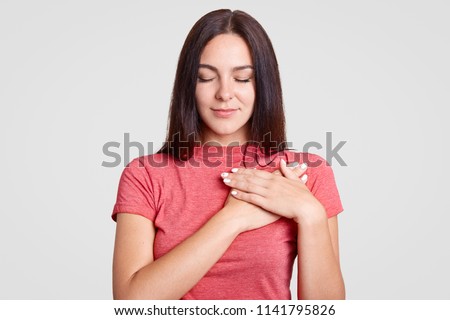 Calm brunette female with closed eyes, keeps both palms on heart, feels gratitude, being touched by something, dressed in casual pink t shirt, isolated over white background. Body language concept