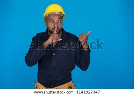 Handsome repairman wearing a navy uniform and helmet, putting his finger in front of his mouth doing shh sign and pointing by his thumb, standing on a blue background.