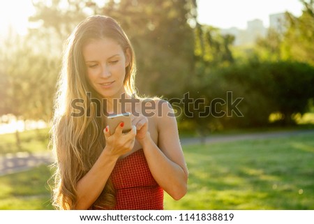 model woman long-haired in red dress holds phone and talking typing text with girlfriends in hand rays of sun background . enamored girl chating with friends in the park