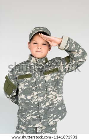 portrait of little kid in military uniform saluting on grey background