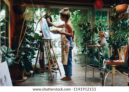 Indoor shot of professional female artist painting on canvas in studio with plants. Royalty-Free Stock Photo #1141912151