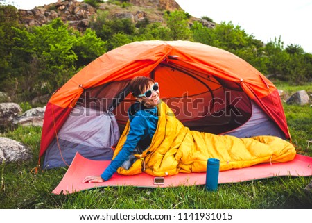 Camping in the forest. A girl is preparing coffee on a geyser coffee machine. A woman sits near a tent in a sleeping bag and prepares coffee against the background of the mountains. Royalty-Free Stock Photo #1141931015