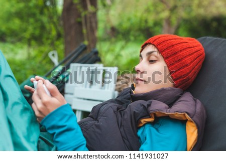 A girl in nature looks into a smartphone. A woman is sitting in a folding chair in the woods and reading from a mobile phone. The brunette sends a message.