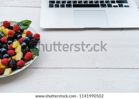 Workspace with laptop and healthy berries in rainbow colours, pineapple, blackberries, blueberries and raspberries with mint on white table. Copy space